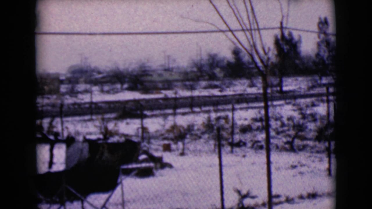 1968 8mm frame from Clarksdale, Arizona showing a snowy roadside neighborhood with fences, bare trees, wires, and distant low buildings.