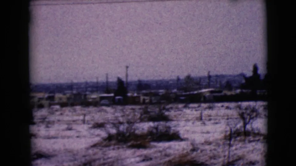 1968 8mm frame of the Clarksdale, Arizona landscape with snow, scattered brush, distant low buildings, poles, and hazy hills.