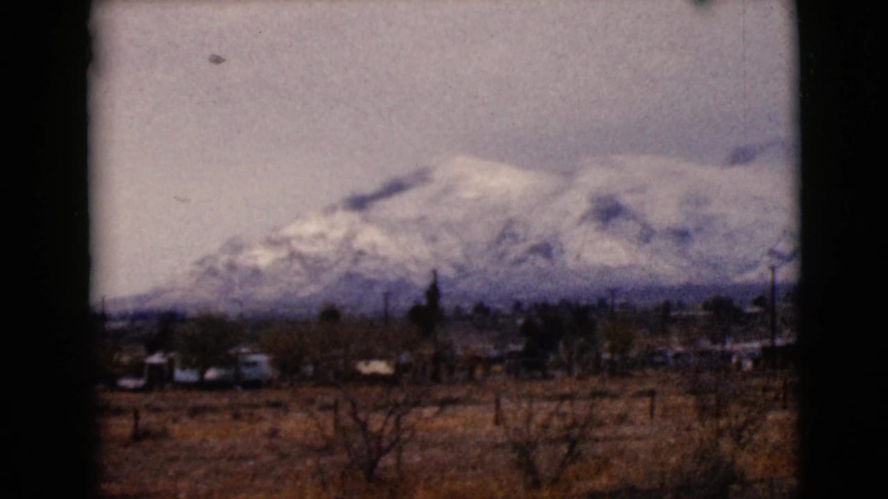1968 Arizona home-movie frame showing snowy mountains beyond dry foreground land, scattered trees, and small buildings.