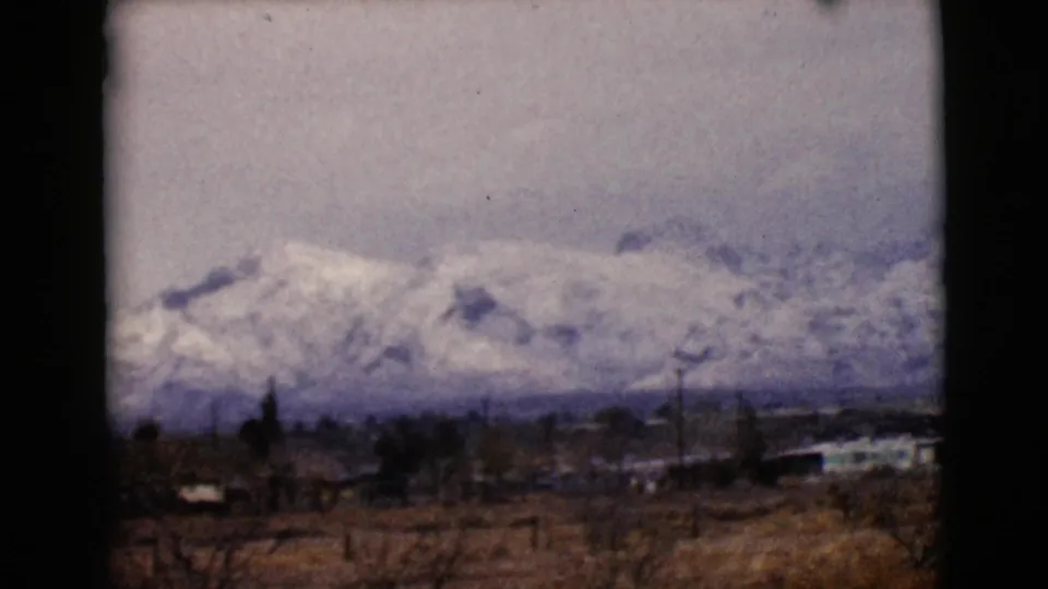1968 Arizona 8mm frame with a broad snowy mountain range above dry fields, utility poles, and low settlement structures.