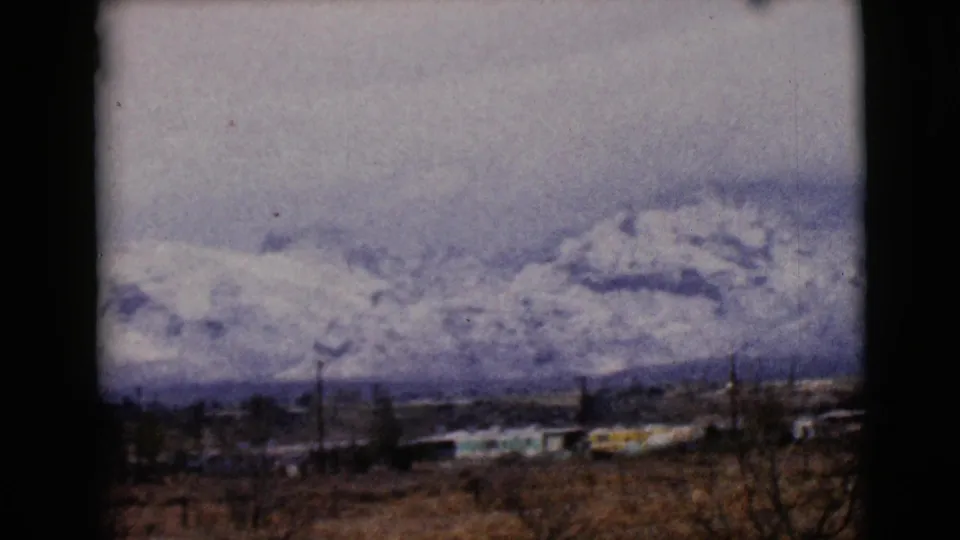 1968 Arizona home-movie frame showing snowy peaks across the horizon, with dry brush, trees, and low buildings in front.
