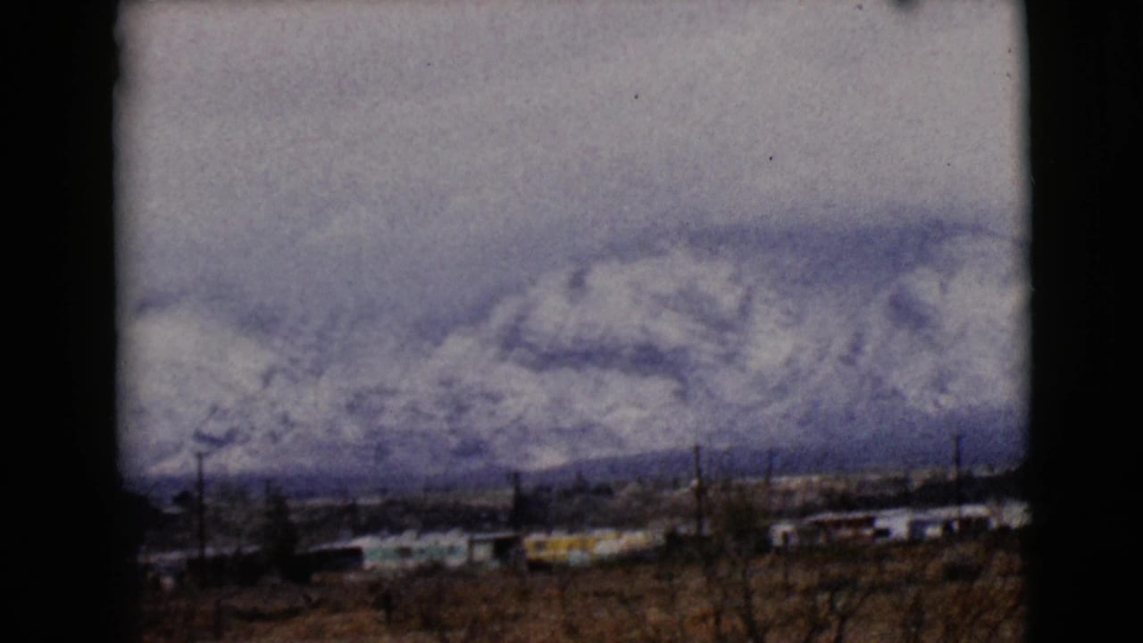 1968 8mm view near Clarksdale, Arizona, showing snowy mountains beyond low rural buildings, bare trees, and utility poles.