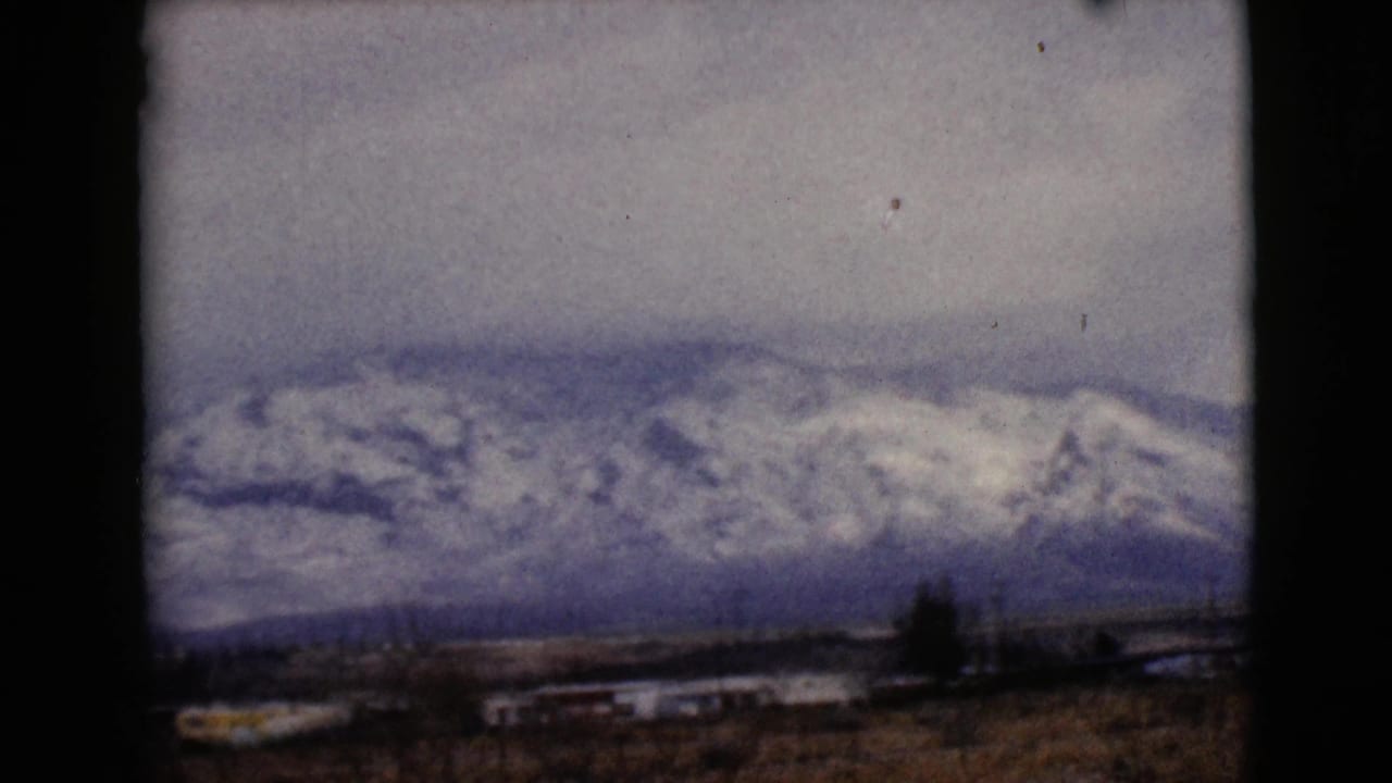 Distant snowy mountain range with low buildings and bare brush in the foreground.
