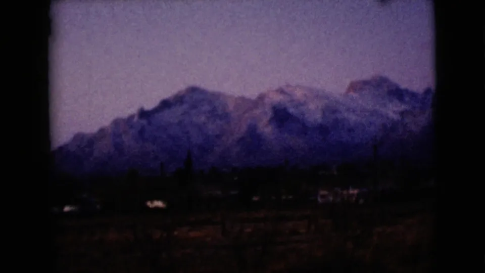 1968 8mm mountain view near Clarksdale, Arizona, showing snow-capped peaks beyond low structures and dark open ground.