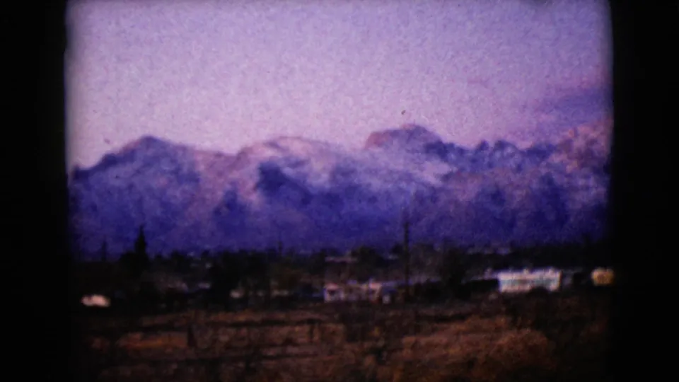 1968 8mm footage near Clarksdale, Arizona, with a snowy mountain range, cloudy sky and low buildings in the foreground.