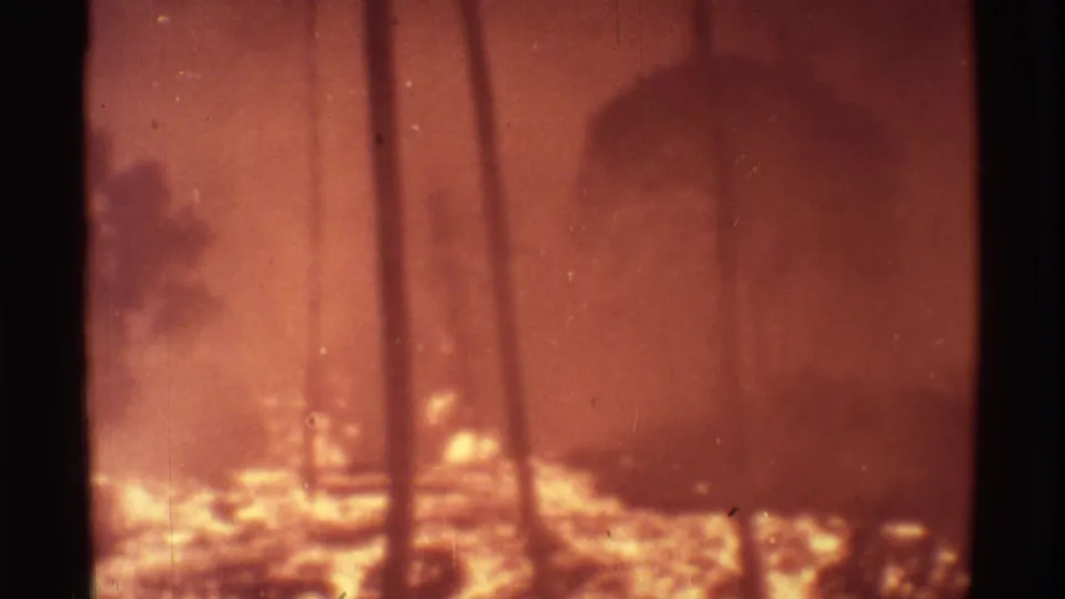 1971 Mauna Ulu 8mm frame showing orange volcanic glow, smoke, and silhouetted palm trunks in soft focus.