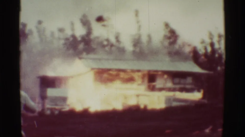 1971 Mauna Ulu 8mm frame showing flames across the front of a low structure, smoke above the roof, and trees in the background.