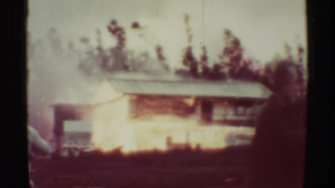 Small structure engulfed by bright flames with smoke and trees behind it.