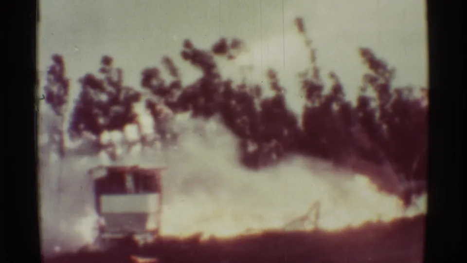 Smoke thickens in this 8mm frame as the structure is partly hidden, with fire visible along the lower edge and dark vegetation above.