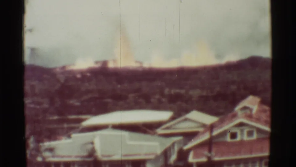1971 Mauna Ulu home-movie frame showing a bright distant fire glow beyond hillside terrain and house roofs.