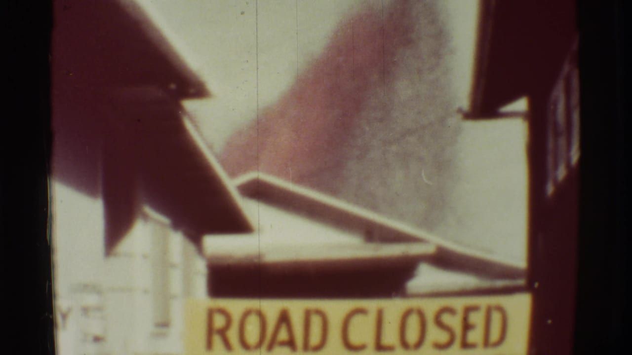 ROAD CLOSED sign below a smoky volcanic plume and roadside structures.