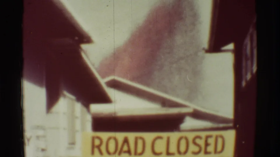 1971 Mauna Ulu 8mm frame showing a ROAD CLOSED sign with smoke or ash rising behind nearby roadside structures.
