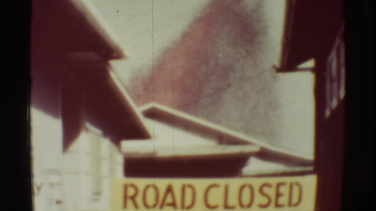 ROAD CLOSED sign with dark plume rising behind roadside buildings.