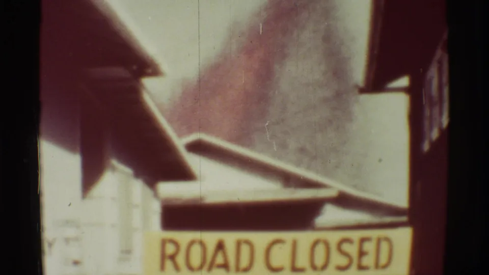 Close 1971 home-movie frame of a ROAD CLOSED sign with smoke, rooflines, and film wear visible around the frame.