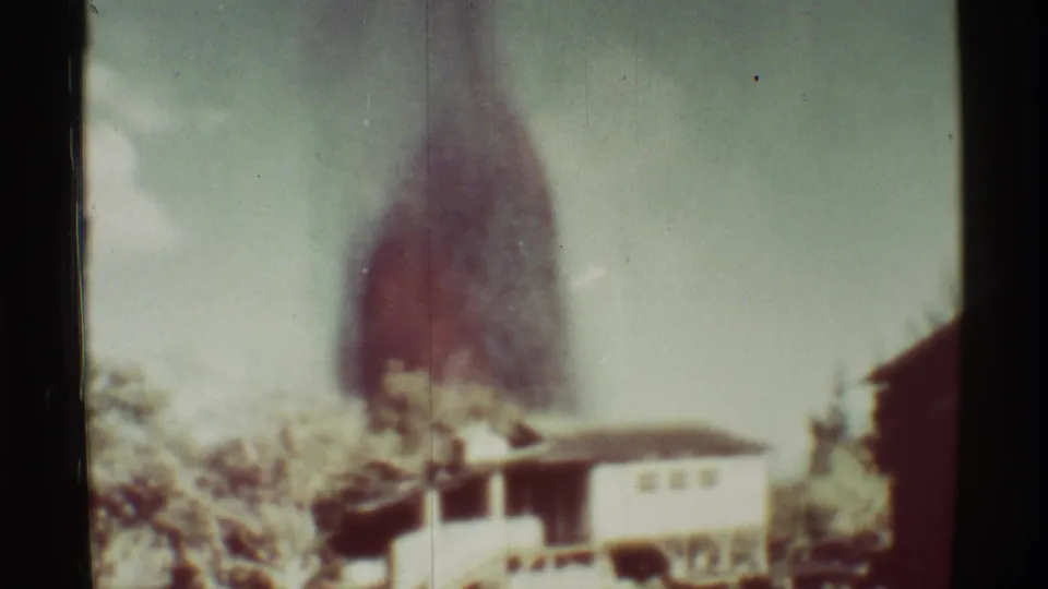 Wider 8mm frame at Mauna Ulu showing a dark plume beyond greenery and a small building near the road.