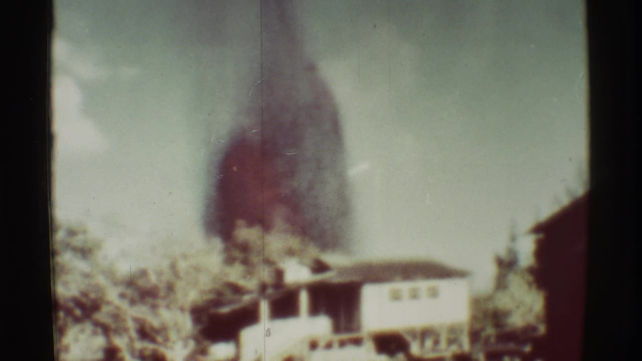 Distant low building beneath a rising dark red smoke plume in daylight.