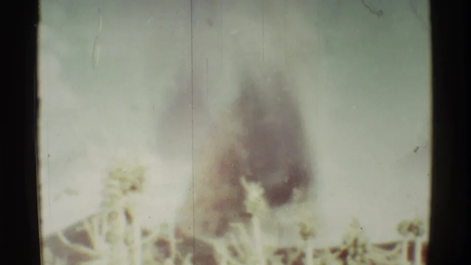 Soft 8mm frame from Mauna Ulu, Hawaii, with heavy smoke and a faint flame glow near trees and a low structure.
