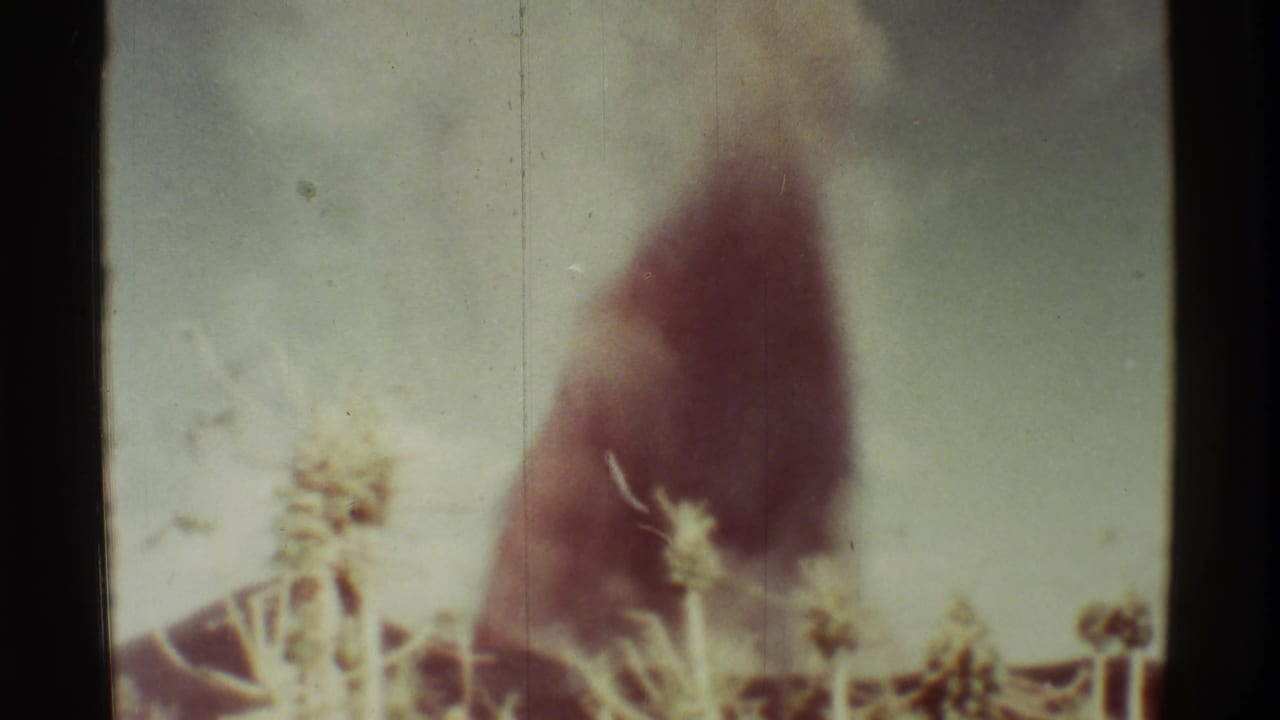 Dark smoke rises behind pale dry vegetation in a faded 8mm frame.