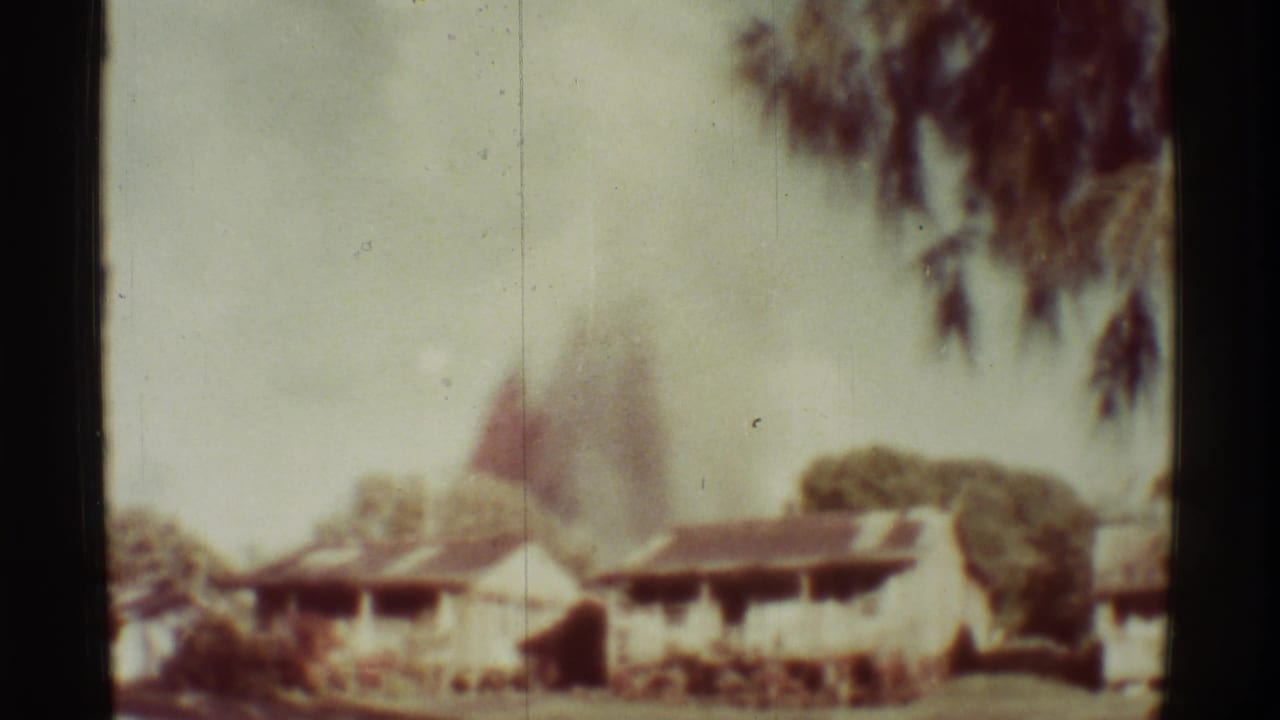 Low buildings and trees with a smoky burst rising in the distance.