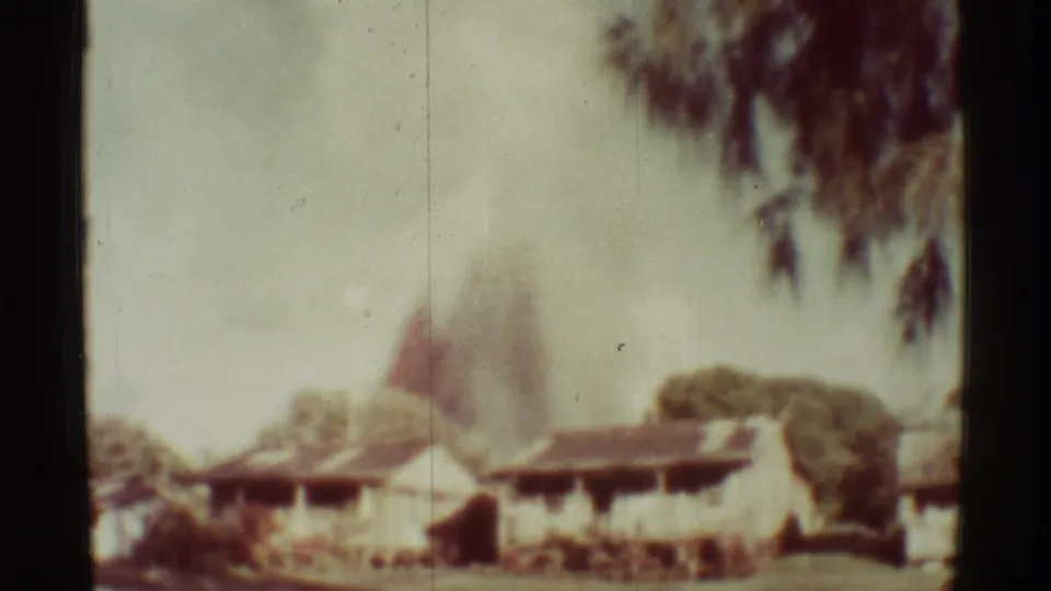 Mauna Ulu, Hawaii frame with low buildings, tree cover, and a pale smoke column with a reddish flare behind the roofs.