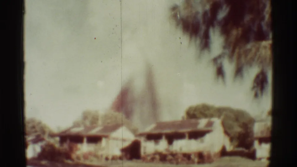 Mauna Ulu, Hawaii 8mm still with low buildings, trees, and a soft smoke column in the distance.