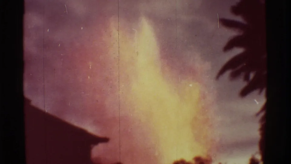 1971 Mauna Ulu eruption frame showing a bright lava fountain behind dark roof and palm silhouettes in faded 8mm color.