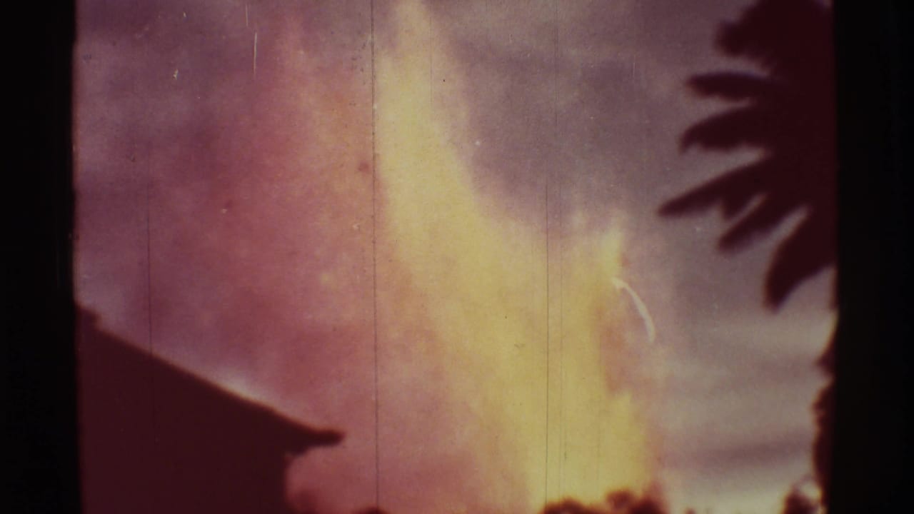 Bright lava fountain rises behind a dark roofline and silhouetted foliage.