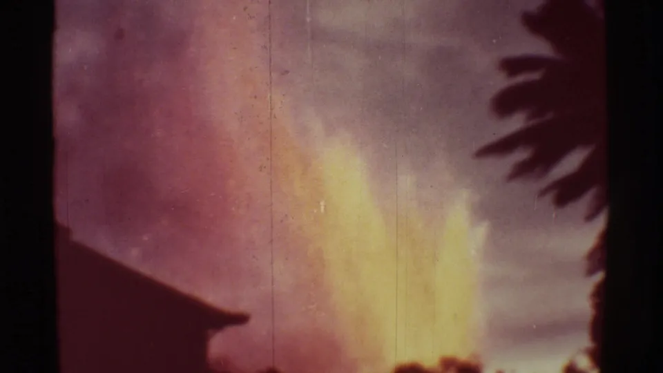 Mauna Ulu eruption frame with a bright yellow-orange plume, faded sky, and dark foreground vegetation on 8mm film.
