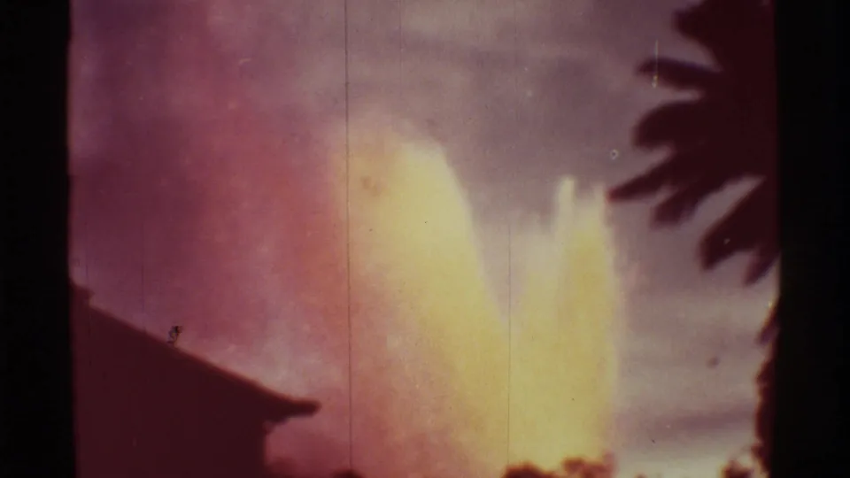 8mm Hawaii volcano frame with a blooming yellow lava fountain, soft purple sky, roofline, and foliage silhouettes.