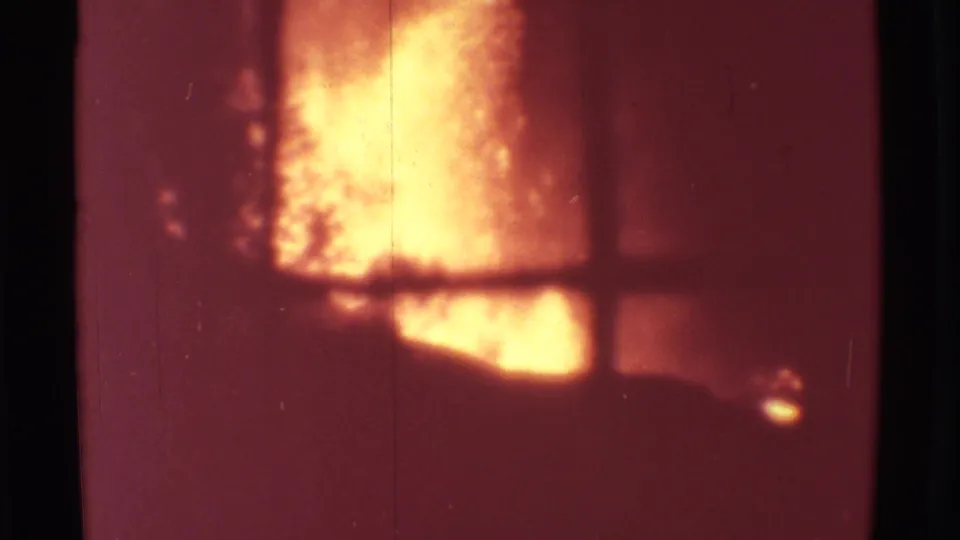 A bright orange-yellow eruption plume at Mauna Ulu is seen through window mullions on grainy 1971 8mm footage.