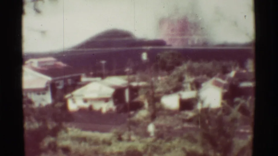 1971 8mm view near Mauna Ulu showing a distant plume behind small buildings and a green hillside.
