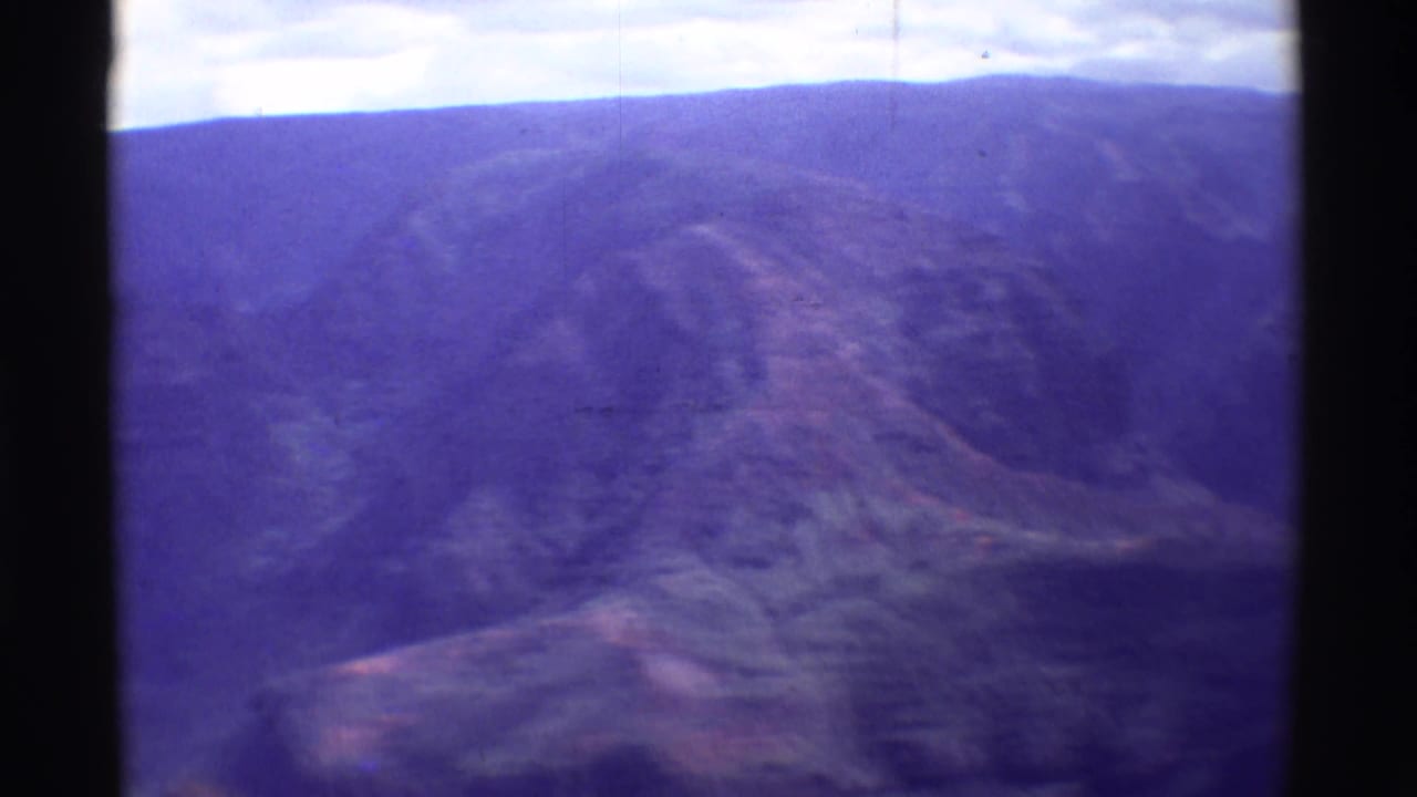 1971 home-movie aerial view over Mauna Ulu, with a broad valley, distant ridge, and softened purple-blue film color.