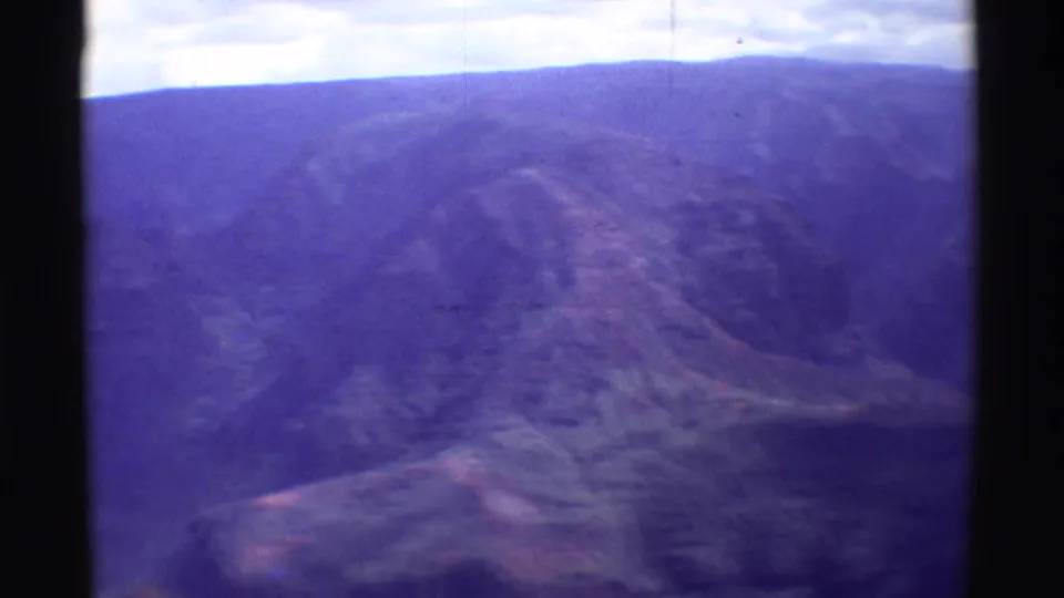 1971 home-movie aerial view over Mauna Ulu, with a broad valley, distant ridge, and softened purple-blue film color.