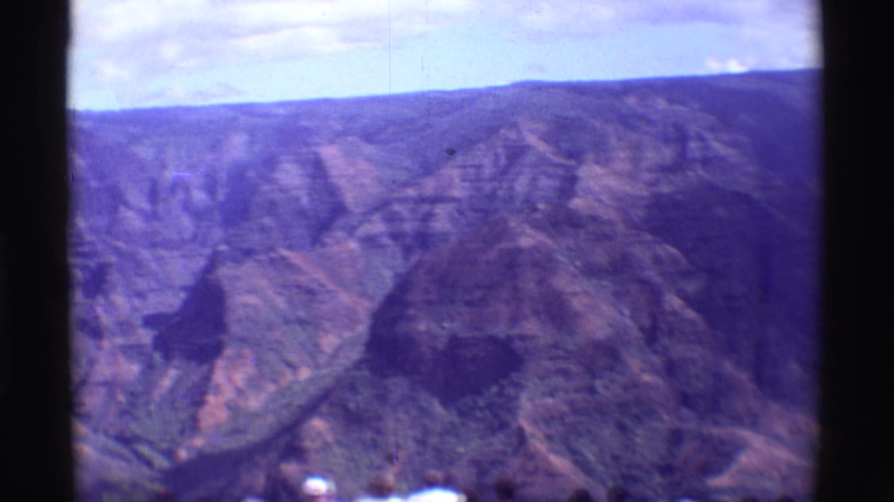1971 8mm Hawaii frame showing eroded canyon ridges, shadowed slopes, a central film border, and small visitors near the bottom edge.