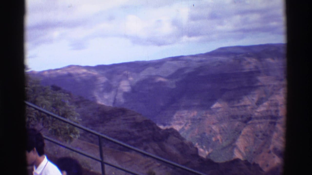 Home-movie frame from the Mauna Ulu, Hawaii metadata location showing an overlook railing, partial visitors, and rugged canyon terrain.