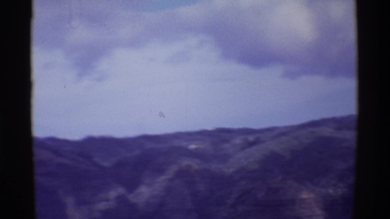 1971 Mauna Ulu lookout view across dark volcanic ridges under clouds, with a small distant bright speck near the hills.