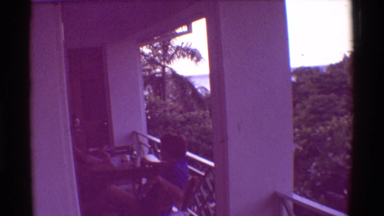 Child seated at a balcony table beside white pillars with tropical foliage and ocean beyond.