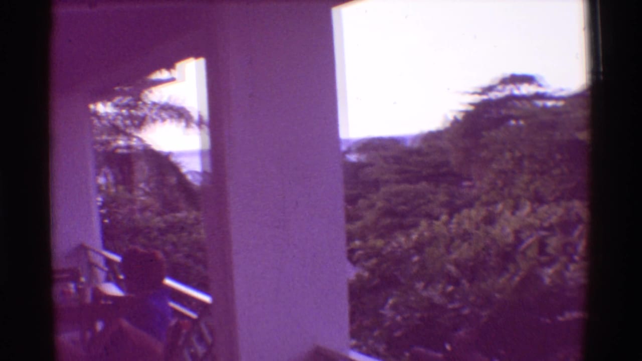 1971 Hawaii 8mm frame looking from a balcony past pillars and railing toward palms and dense tropical greenery with water beyond.