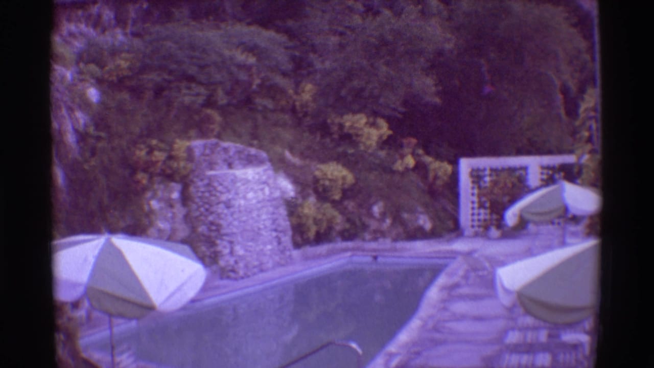 1971 Hawaii home-movie frame showing a curved pool edge, white patio umbrella, stone wall, paved deck, and tropical vegetation.