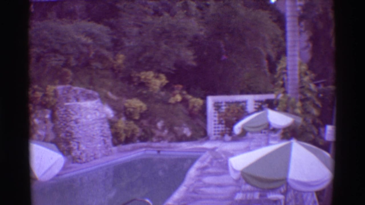 Swimming pool bordered by palms, greenery, stone paths, and striped umbrellas.