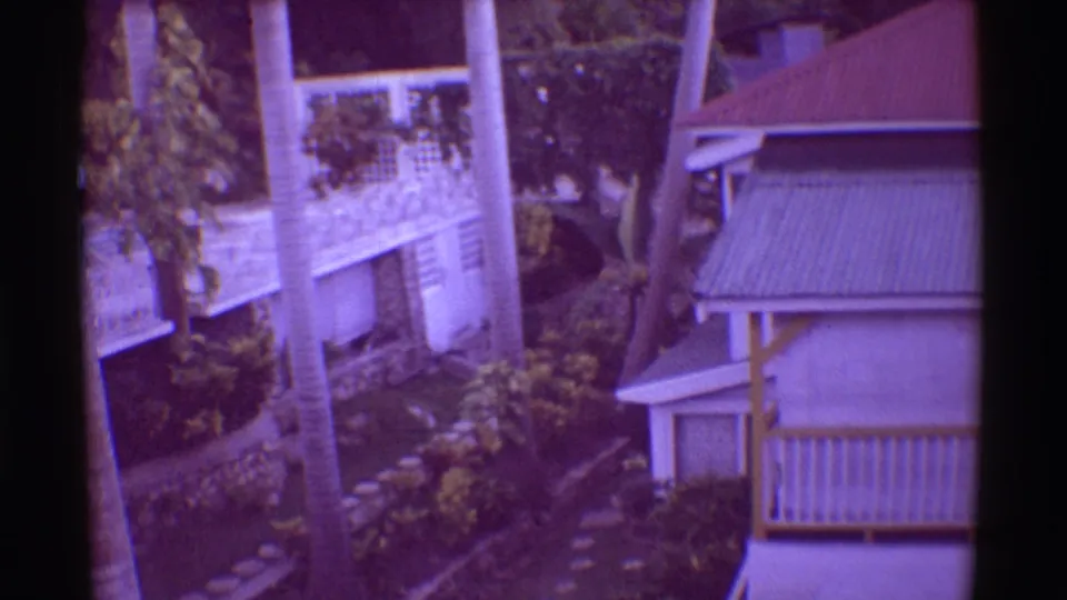 1971 Hawaii 8mm frame showing a resort exterior with white balconies, palm trees, stone edging, and dense tropical planting.