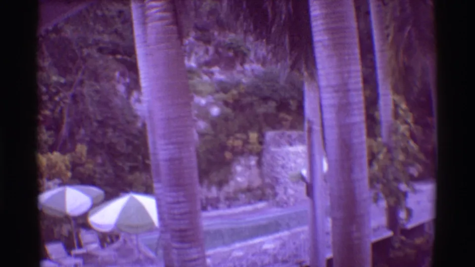 1971 Mauna Ulu, Hawaii 8mm frame showing palm trunks, white umbrellas, stone landscaping, and lush resort greenery.