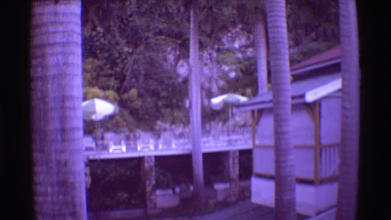 1971 Mauna Ulu, Hawaii 8mm frame with a resort deck, white umbrellas, palm trunks, railings, lush foliage, and a building edge.