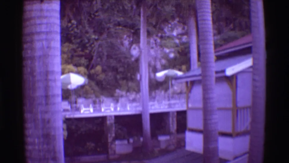 1971 Mauna Ulu, Hawaii 8mm frame with a resort deck, white umbrellas, palm trunks, railings, lush foliage, and a building edge.