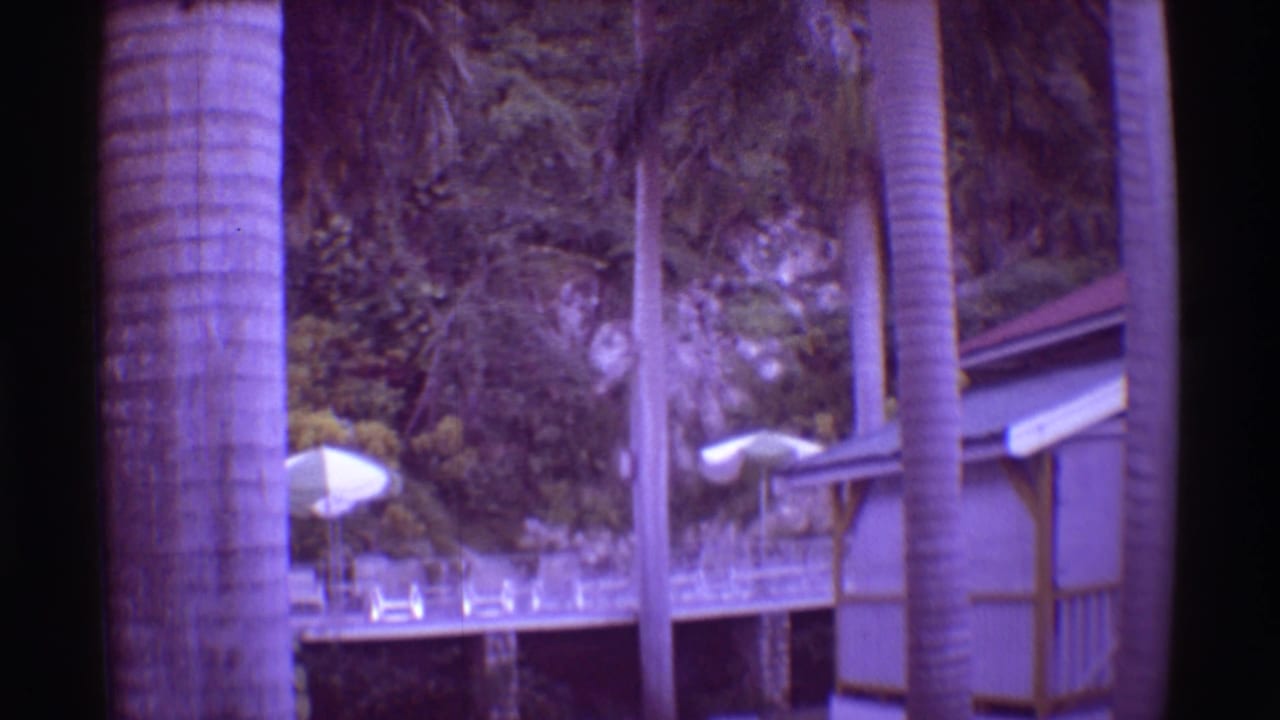 1971 Mauna Ulu, Hawaii frame showing palm trunks, a raised resort deck, white umbrellas, chairs, railings, and dense tropical foliage.