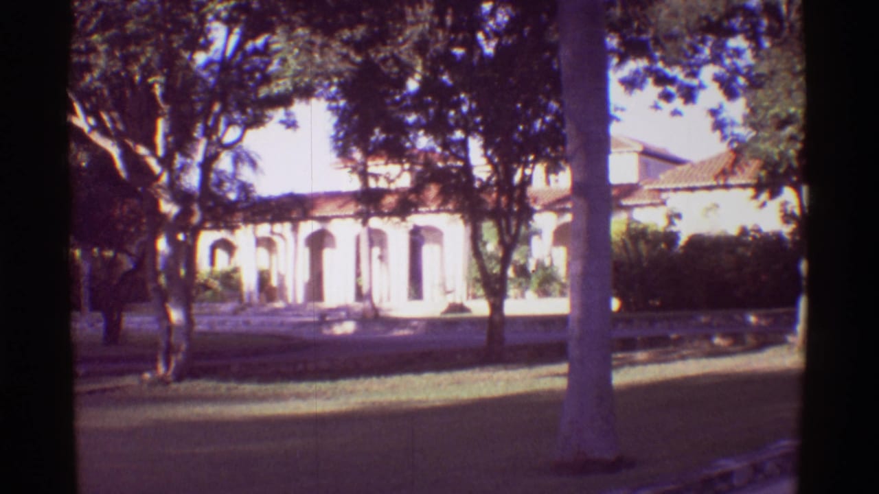 1971 Mauna Ulu, Hawaii frame showing an arched resort facade behind trees, with lawn, shadows, and a palm trunk in the foreground.