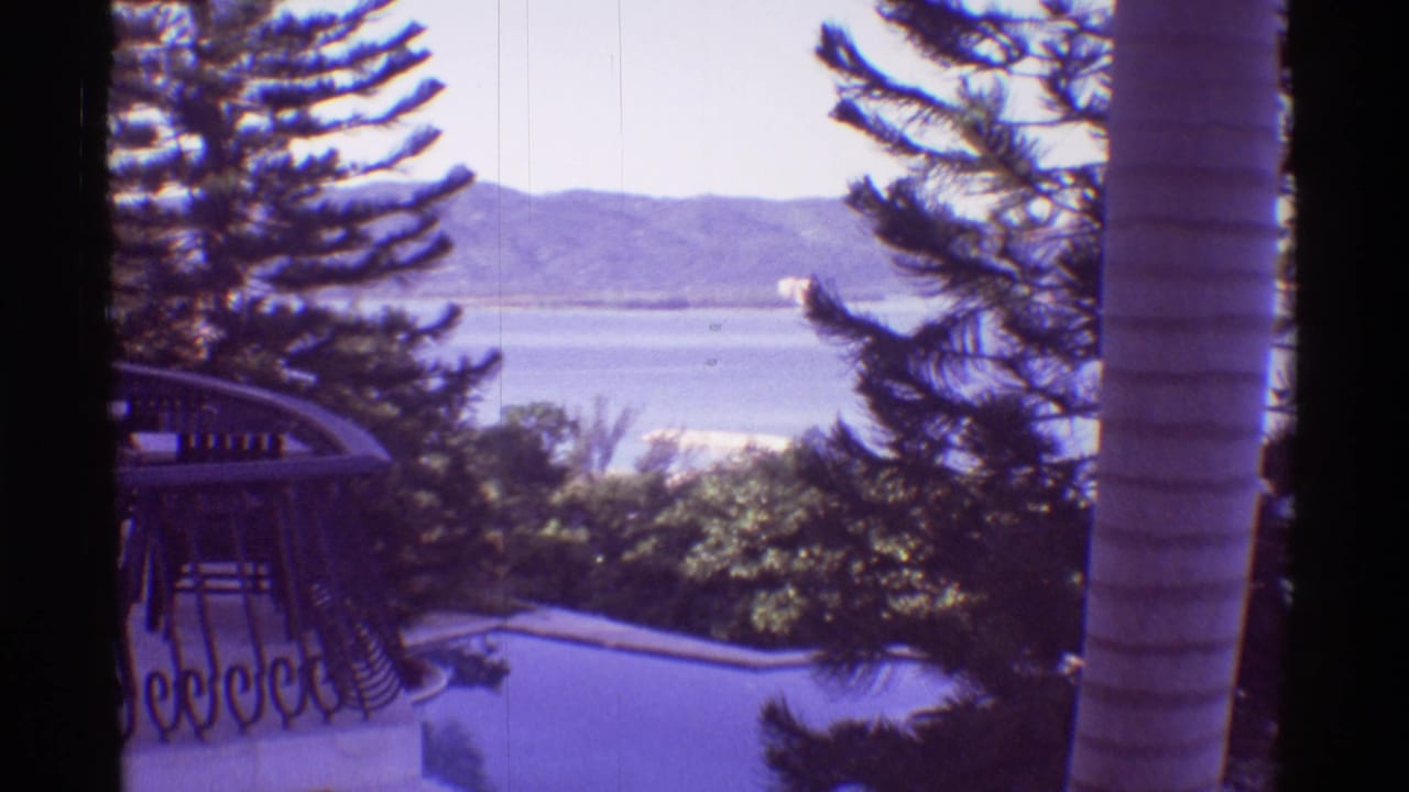 1971 Hawaii balcony view with an ornate railing, swimming pool, palm trunk, broad water, and distant mountains.
