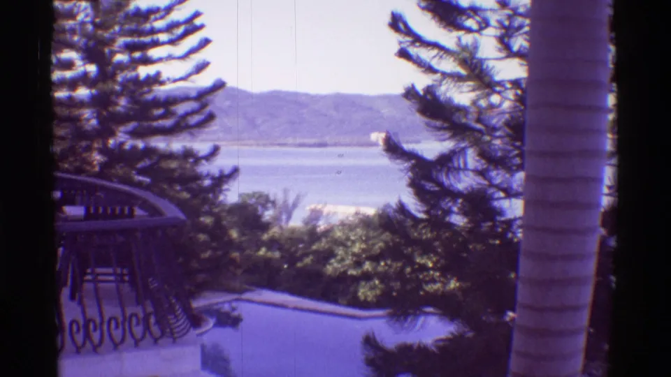 1971 Hawaii balcony view with an ornate railing, swimming pool, palm trunk, broad water, and distant mountains.