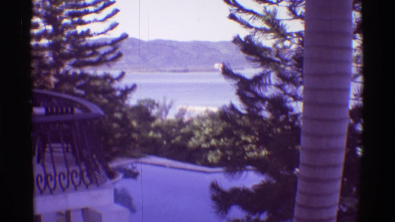 1971 Hawaii home-movie frame looking past a pool and palms toward open water and far mountains.