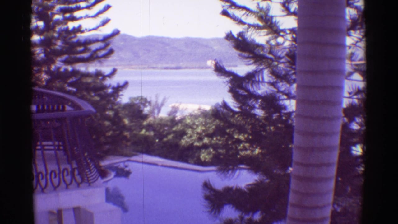 Balcony view of a pool, palm trees, ocean, and distant mountains.